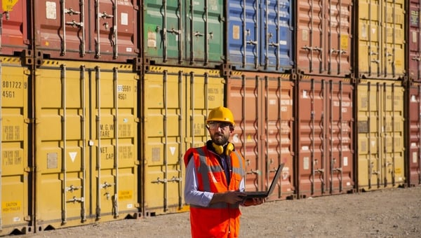 man holding a laptop standing in front of shipping containers 