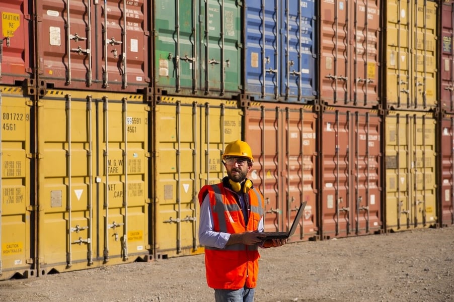 man holding a laptop standing in front of shipping containers 